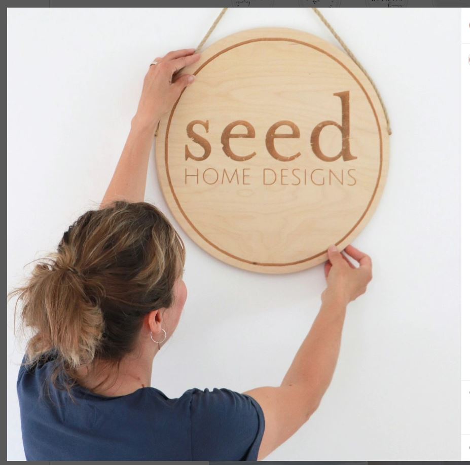 Naomi Sisson, founder of Seed Home Designs, proudly hangs a sign bearing the company's name, embodying her commitment to sustainable living and unique design.